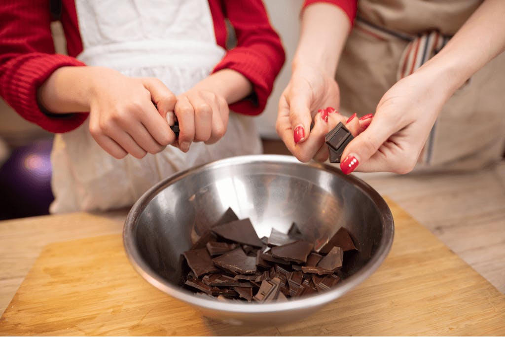 A woman and a younger woman break chocolate into a metal bowl so they can make Valentine's Day Chocolate.