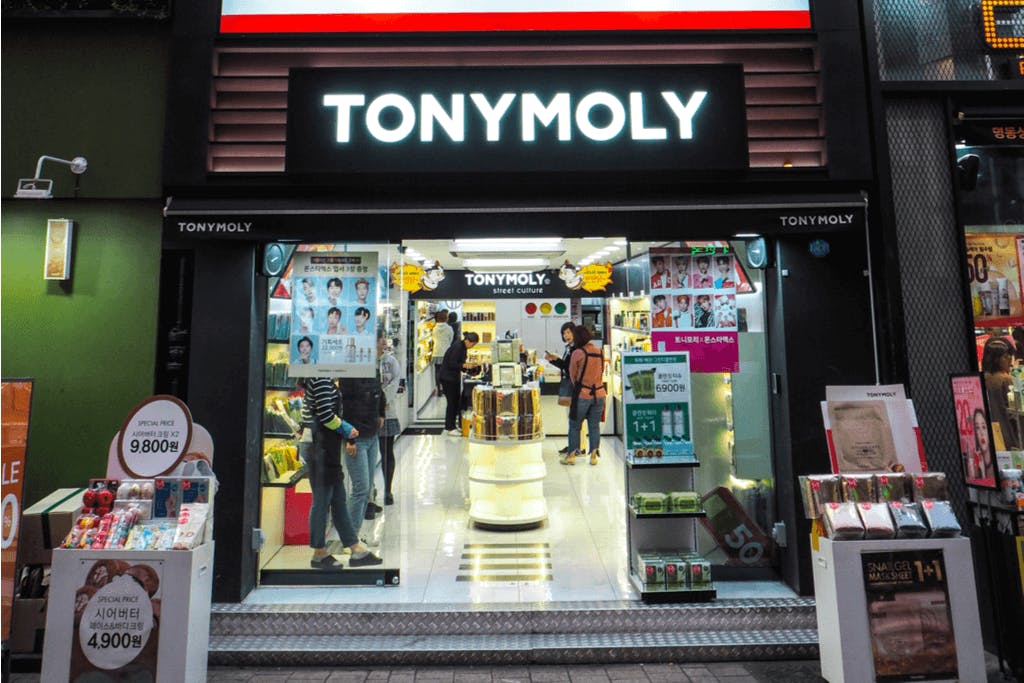 The storefront of TONYMOLY, a K-beauty brand popular for Korean lip gloss, tint, and more, with some people inside shopping and some products on display in front.