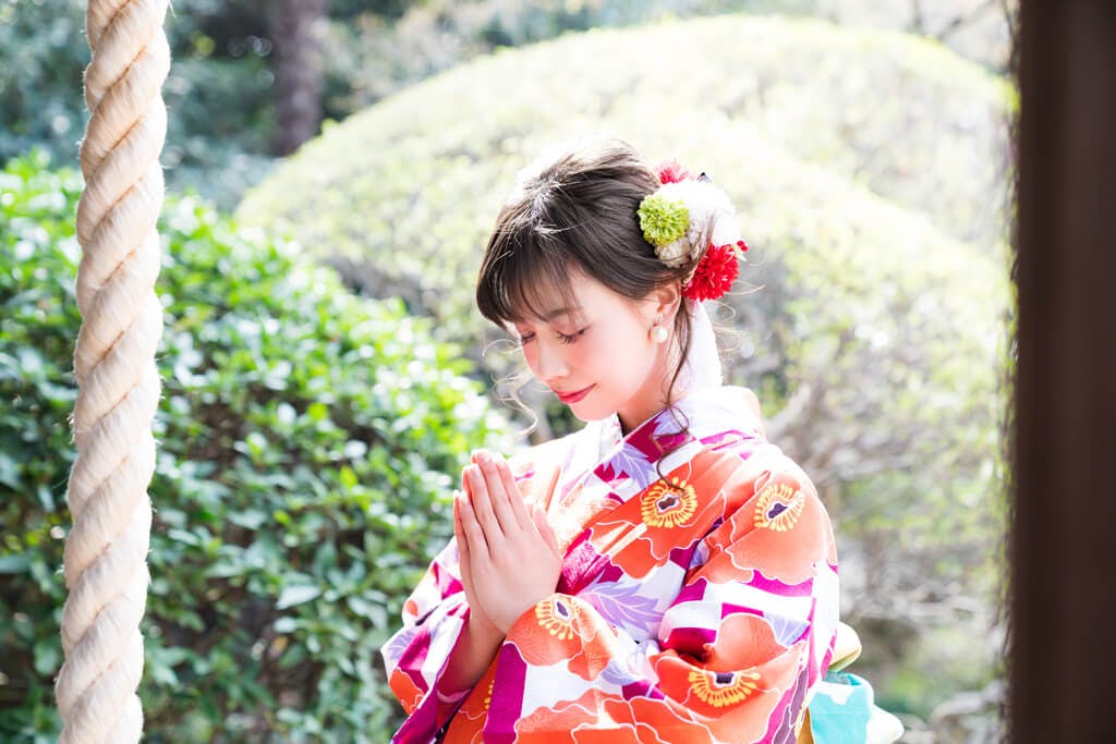 A young woman in a kimono participates in the New Year prayer (hatsumode) with a nature scene behind her