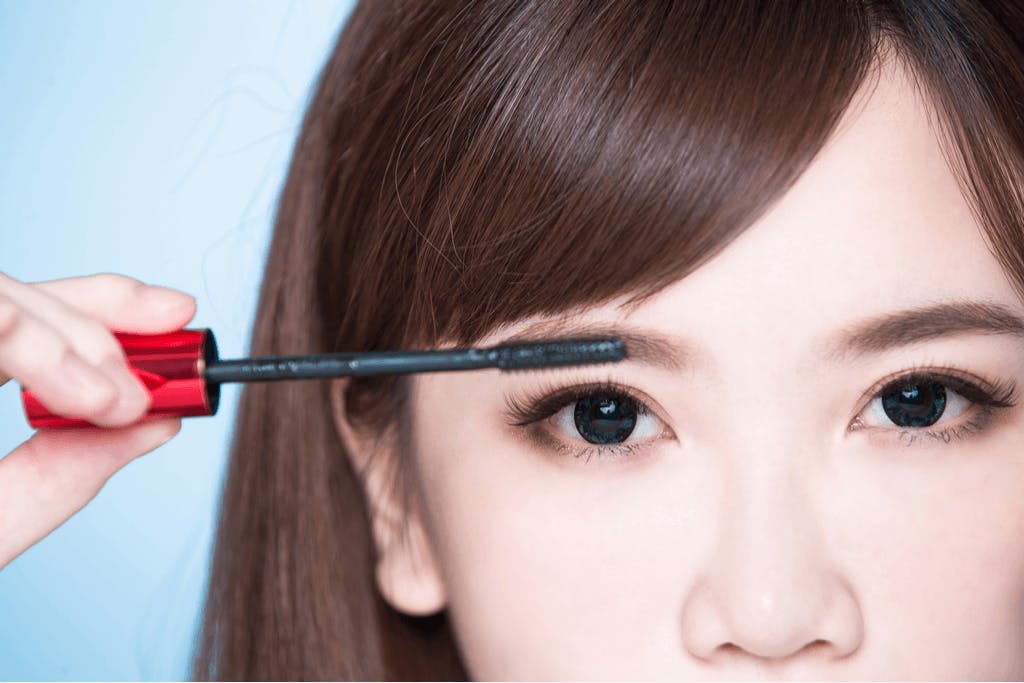 A woman applies a black mascara to finish her eye makeup which uses colors common in current Japanese makeup trends.