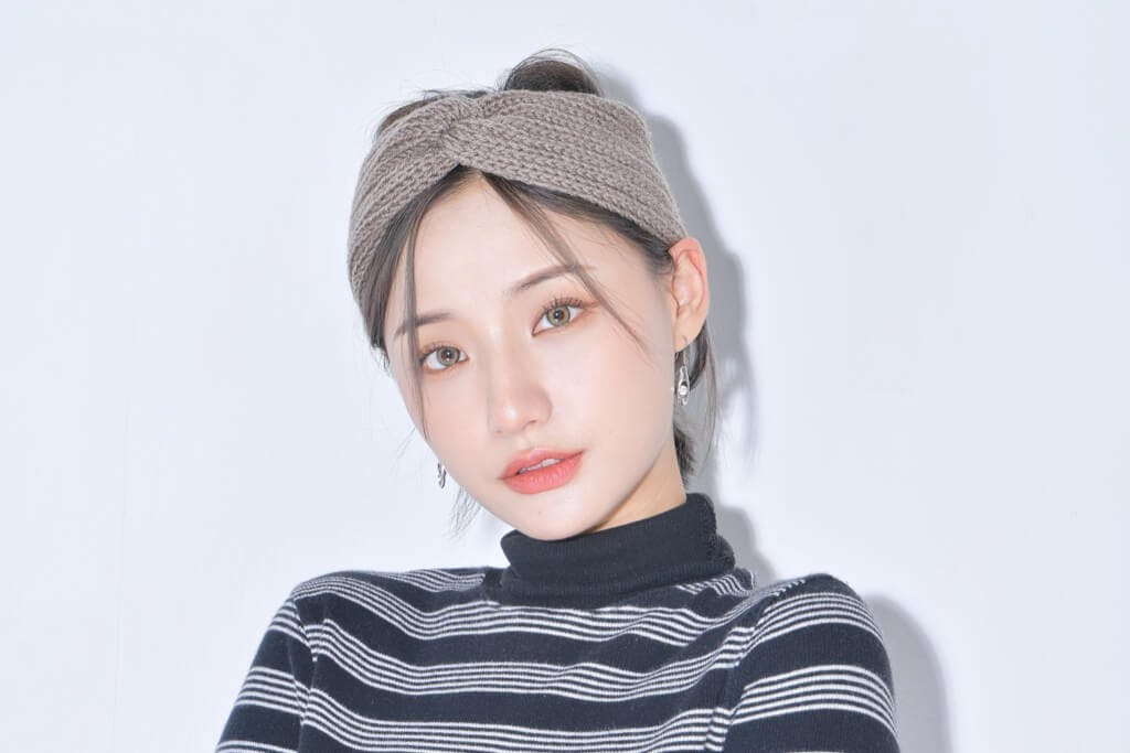 A young woman in black and white sweater and headwrap stands in front of a white background after applying her Peach C makeup.