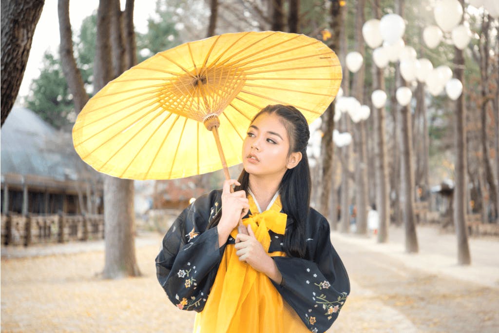 A woman walks through a historical Seoul site in a black and yellow Hanbok and yellow parasol with a face of modern makeup. 