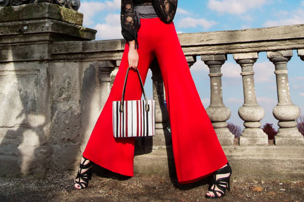A woman stands in front of part of a bridge wearing red wide leg pants while holding a white, black and red bag and wearing high heels