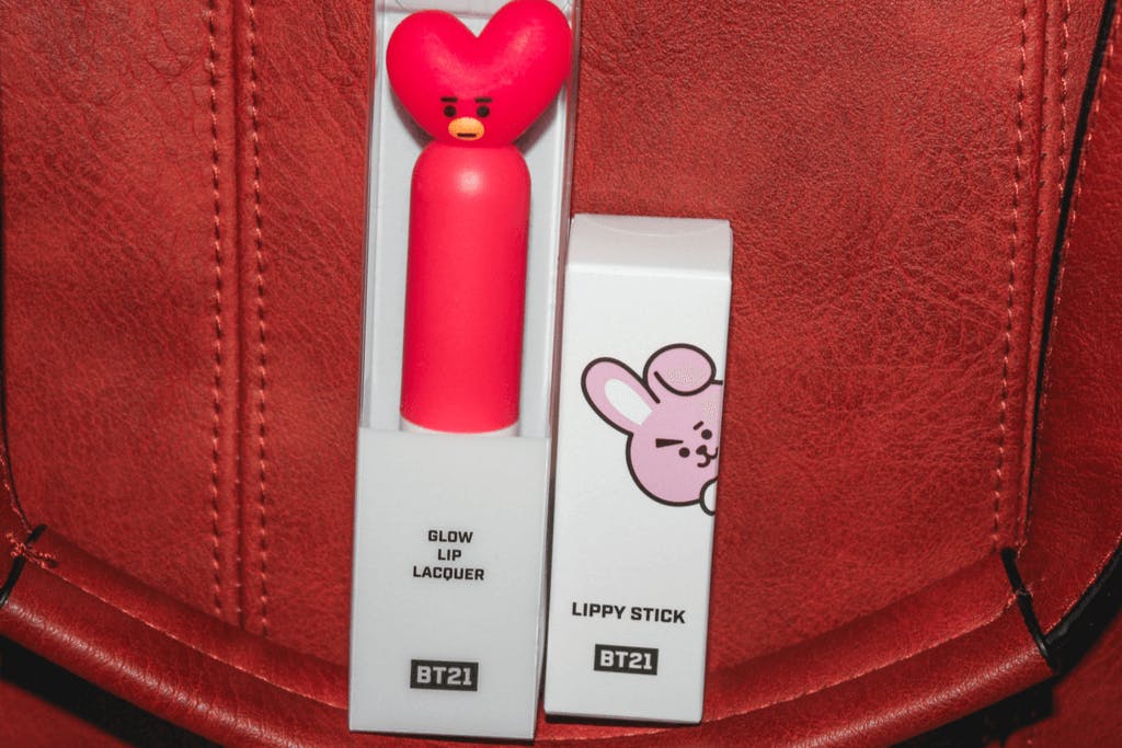 Two BT21 makeup products, a lip lacquer featuring Tata and a lip tint featuring Cooky, with a red bag as the background.