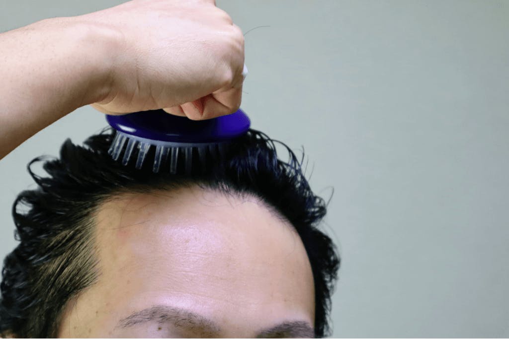 A man uses a blue scalp massager with white prongs on his head to massage his scalp, a common step in the Korean hair care routine.