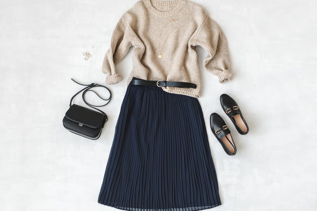 A Korean Fall fashion look made up of a midi skirt, knit sweater, black purse, black shoes, and black belt laid on a marble background