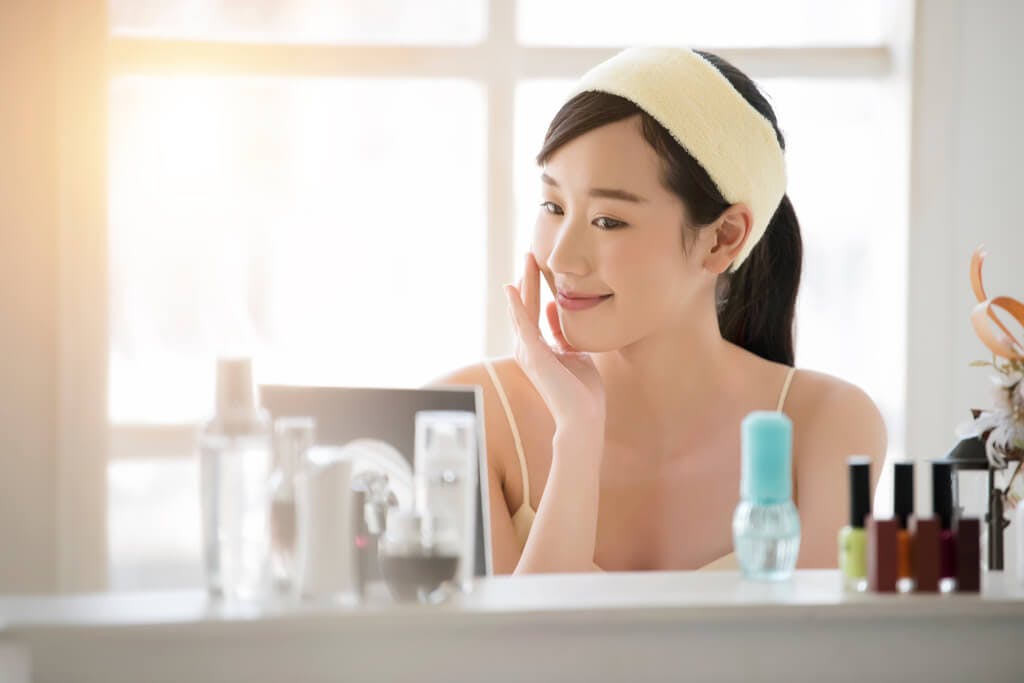 A woman touches her skin with korean beauty products in front of her as she looks in the mirror