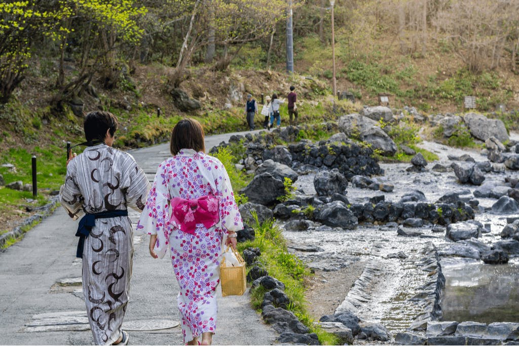 A couple in traditional Japanese clothing walks towards an onsen with rivers of warm water streaming by.