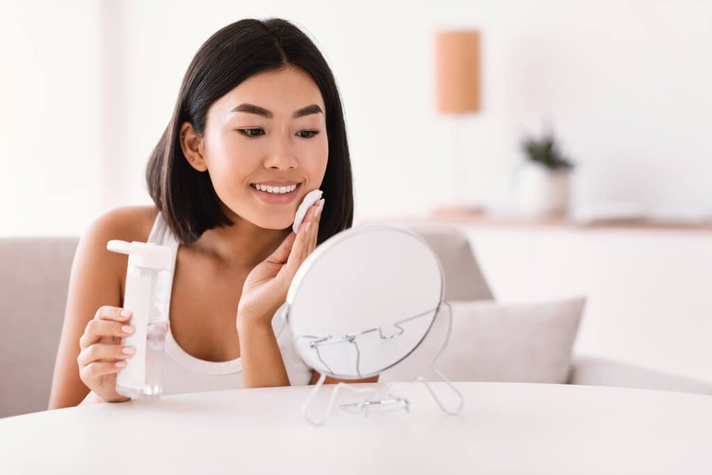 A woman sits in her white living room in front of a white mirror while putting on toner