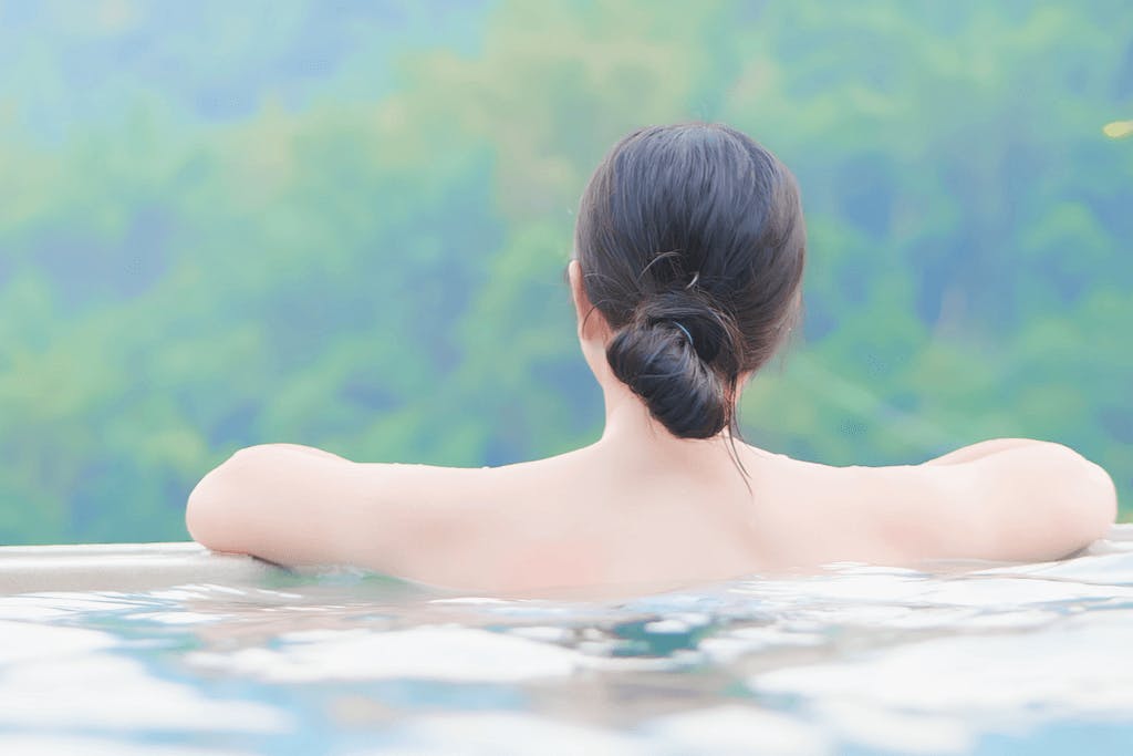 A woman soaks in an outdoor onsen as she looks out at the forest in the background.