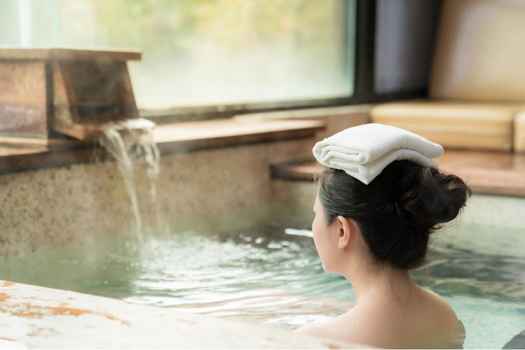 A woman with a towel on her head sits in a sento with a wooden spout pumping in new water
