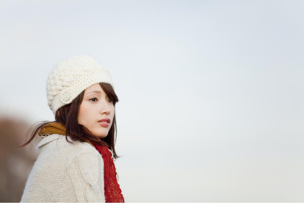 A woman sits and looks into the distance while wearing a knit cap, a scarf, and a cardigan.