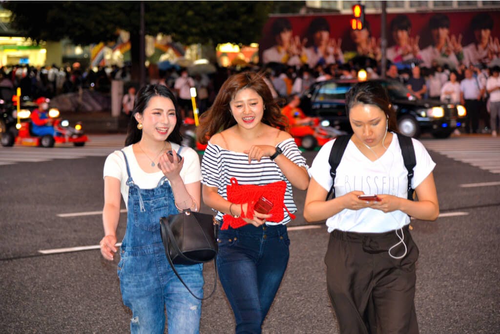 Two young women talking to each other while a third messages on her phone on the streets of Shibuya in front of a Mario Kart tour 