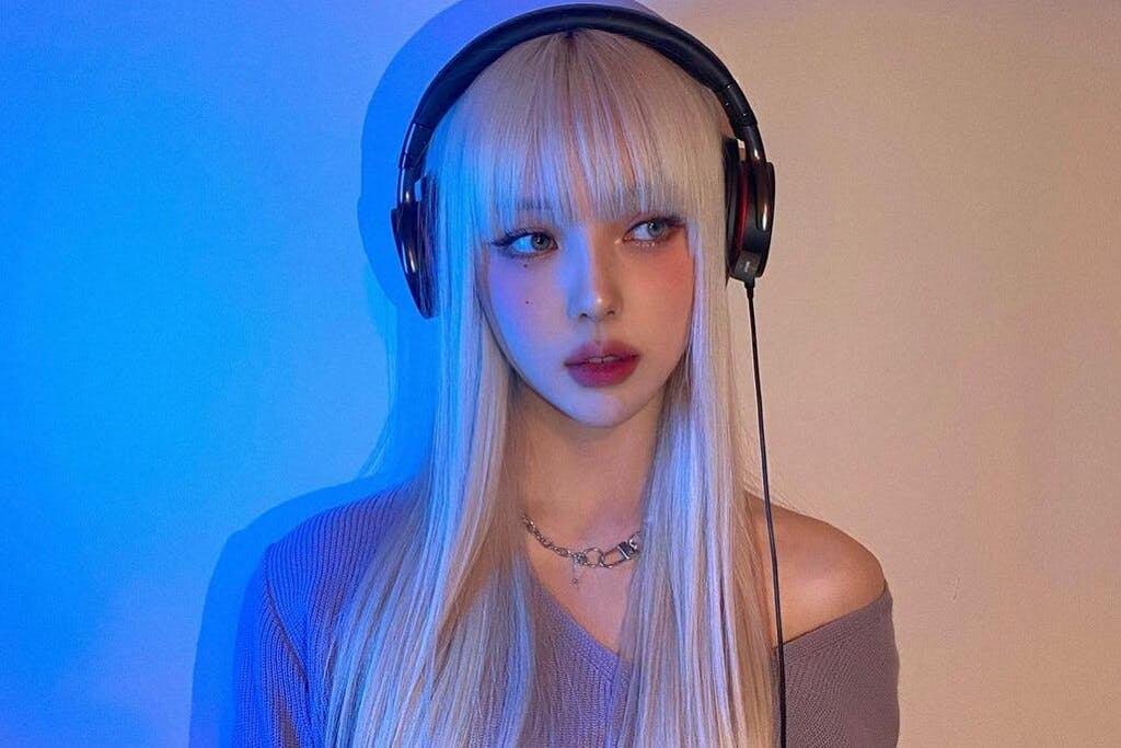 Pony, a popular Korean Instagram influencer and YouTuber, with long blond hair and gorgeous makeup stands in front of a white background wearing headphones.