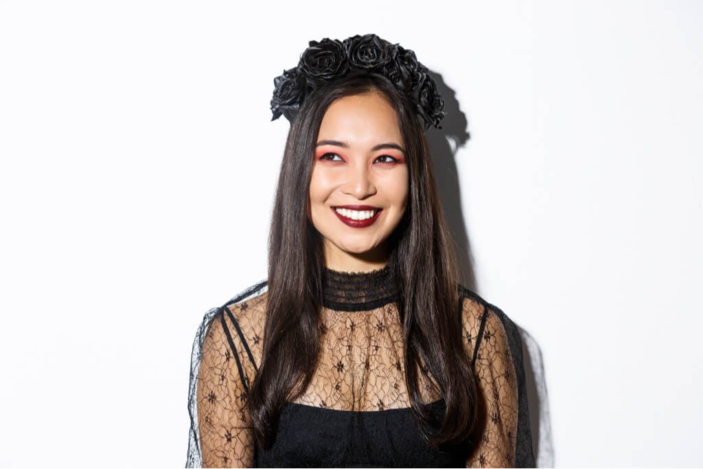 A woman smiles in a black dress with sheer shoulders and sleeves and a black flower crown with purple eyeshadow.