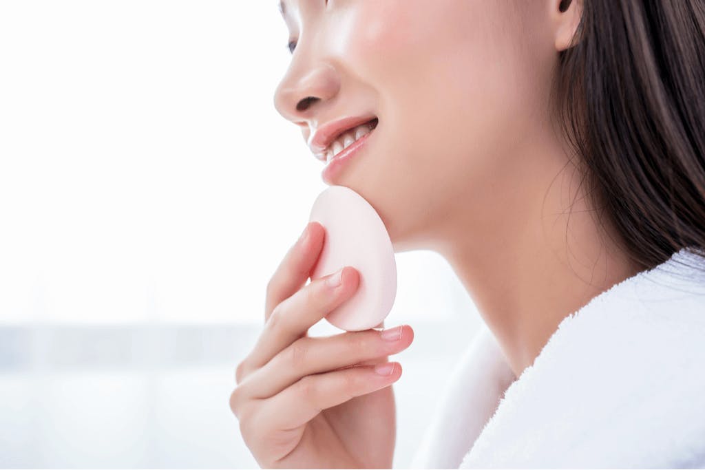 A woman in a white bathrobe blending a light foundation into her face with a blender sponge.