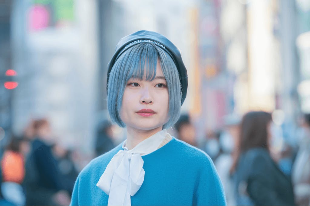 A young woman stands outside in modern Shibuya with beautiful, shiny blue hair, wearing a blue sweater with a white ribbon.