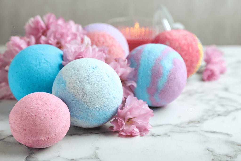 Many different colors, some multi-colored, and sizes of Japanese bath bombs with flowers among flowers and a candle on a marble table.