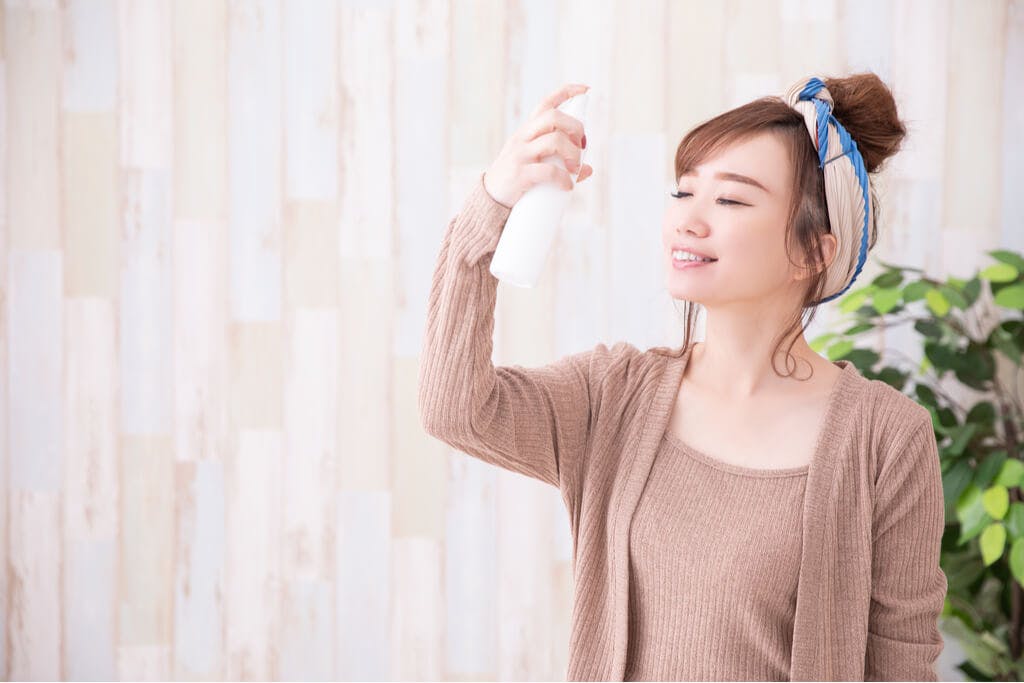 A woman wearing a headwrap sprays a korean product for dry skin on her face while smiling