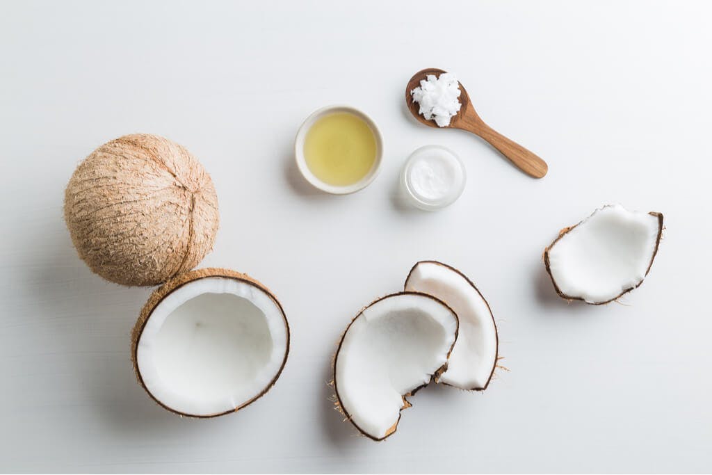 Coconuts sit on a table, most of them cracked open, with an oil and cream next to it and a wooden spoon with coconut shavings