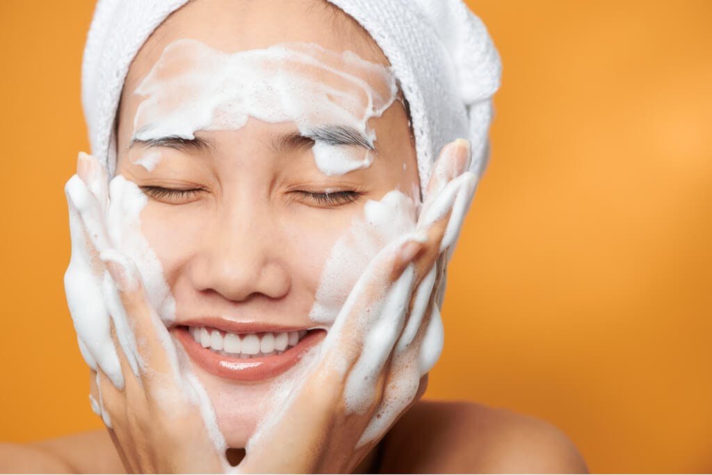 A woman washing her face with a towel wrapped around her face in front of an orange background while smiling