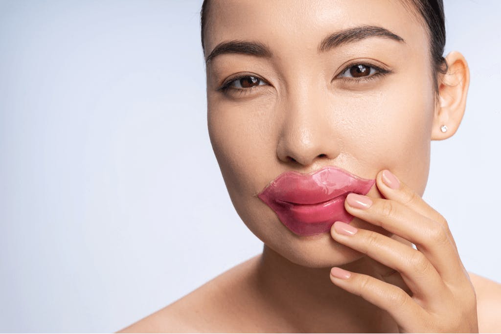 A woman wearing Korean lip mask and pressing it to her face with one of her hands in front of a white background.