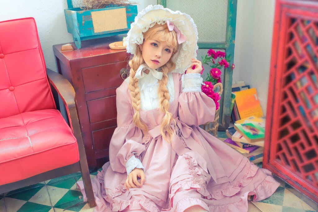A girl in Lolita-style clothing sits in a room next to a chair and in front of a drawer.