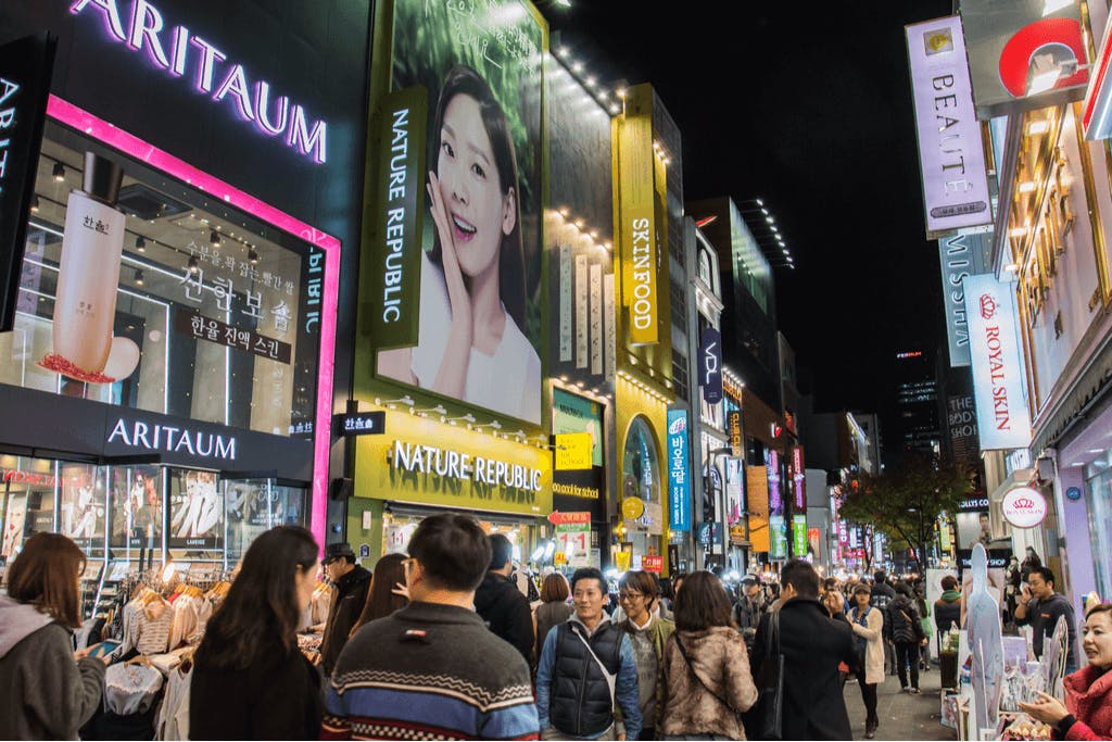 A nighttime picture of a shopping street with many people walking around and with many Korean skincare and Korean hair care brands billboards and signboards lighting up the street.