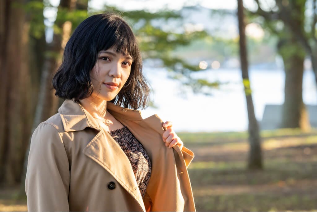 A woman stands in a park wearing Japanese fall fashion like a trench coat while holding it open with one hand