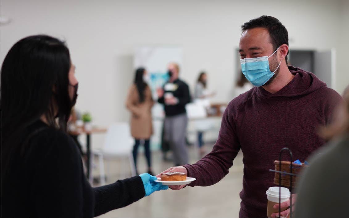 Girl handing a donut on a plate to a guy who is smiling and holding a coffee.