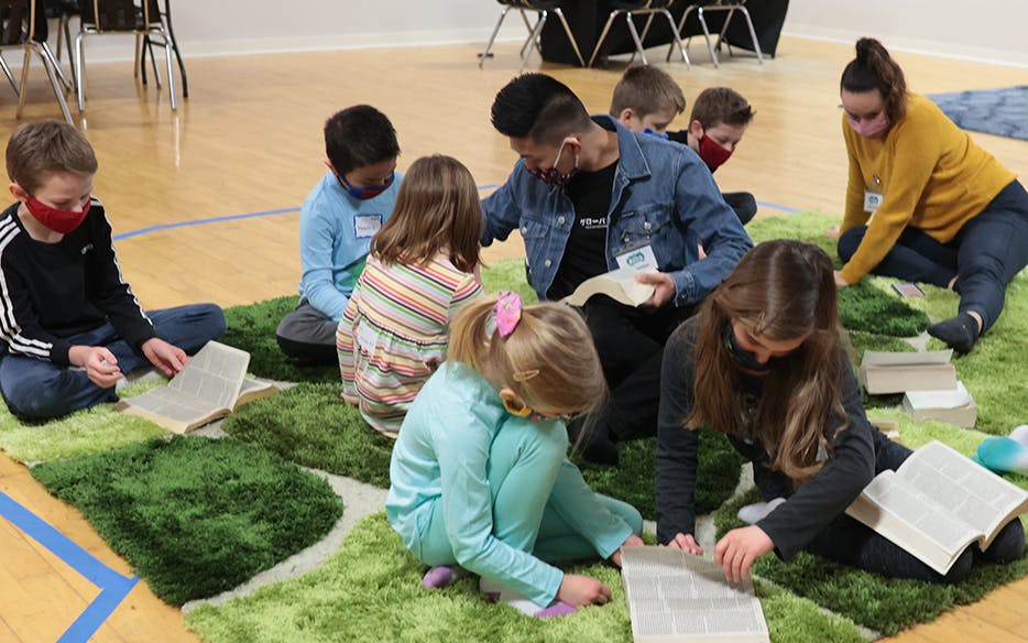 Children surrounding a teacher on a large rug, reading their Bibles.