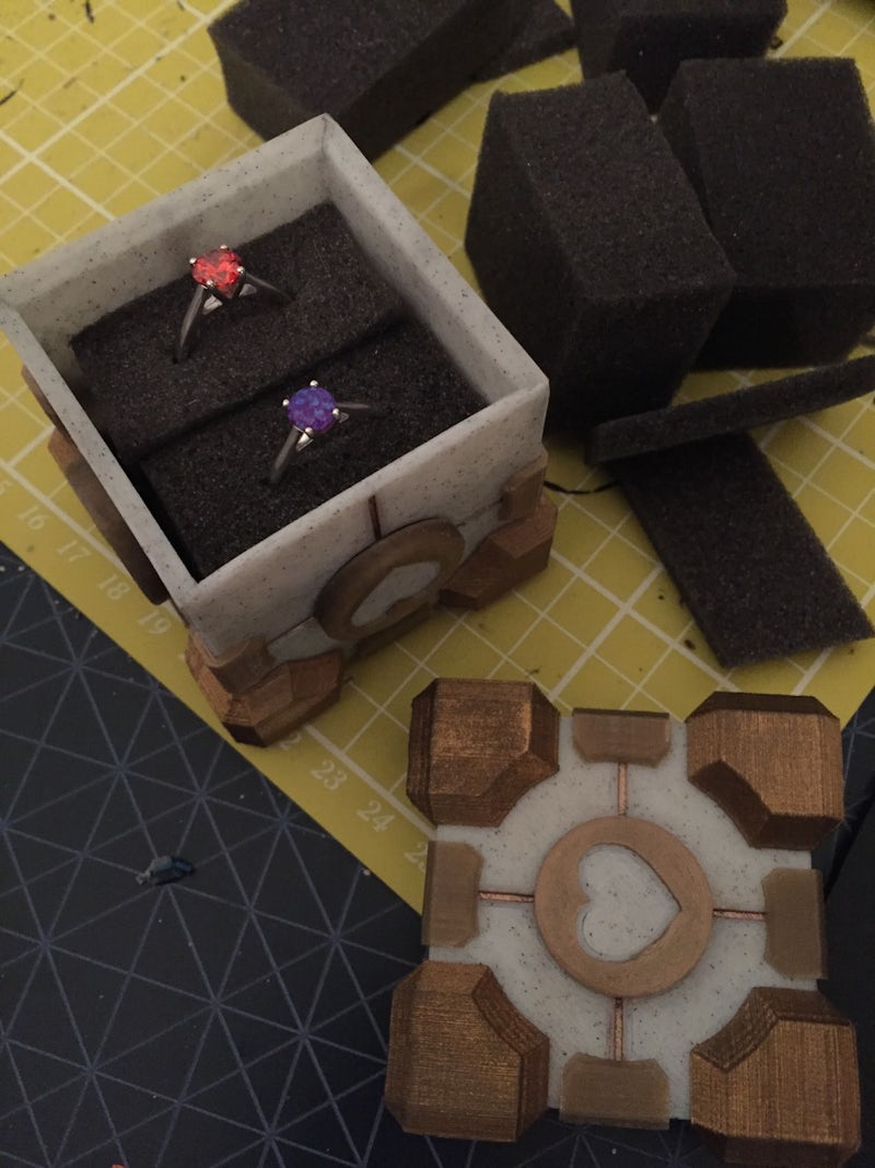 A small wooden box with a heart design, has been place on a table with the lid removed. Inside are two delicate crystal rings, one red the other a rich purple colour.