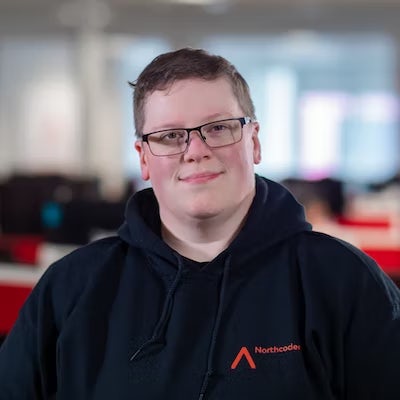 A smiling bespectacled woman with short hair stands centrally in a photograph, she is wearing the iconic Northcoders hoodie.