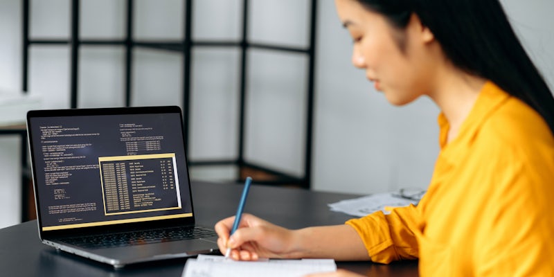 Woman making notes with code on an open laptop next to her