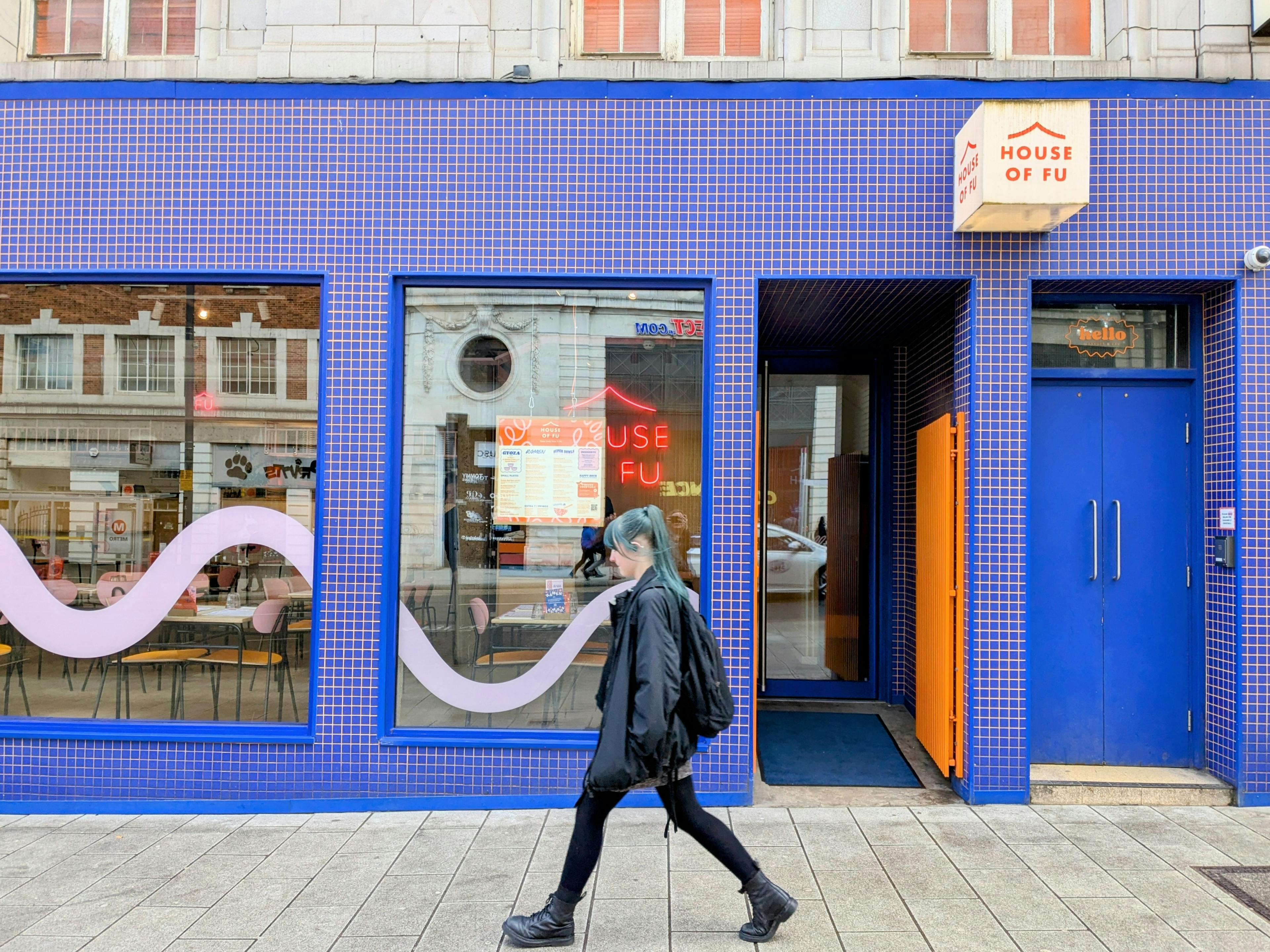 A person in black with blue hair walking past House of Fu, Leeds