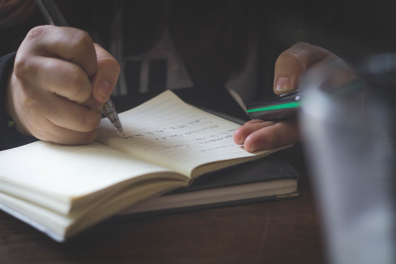 A man writing in a notebook while holding his phone - it is the main image for the article "Best Bullet Journal for iPhone: The Top 9 Bullet Journals Reviewed"