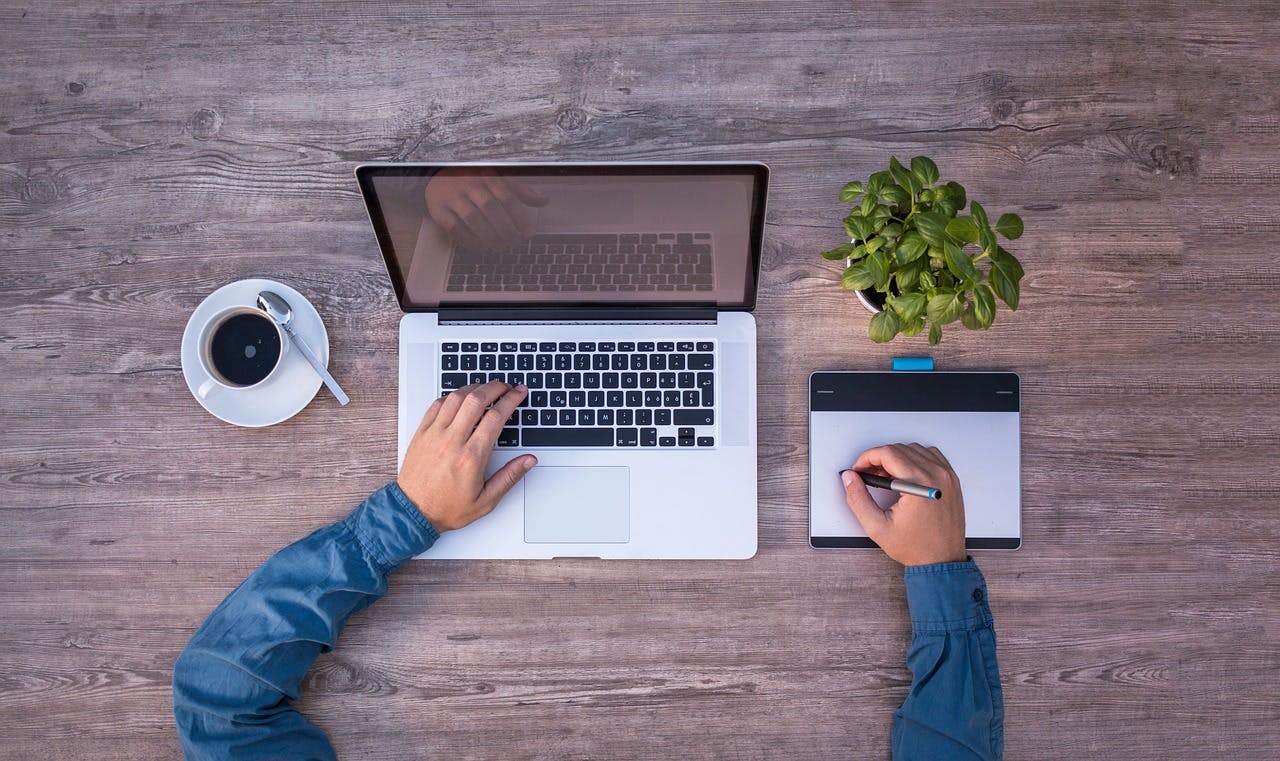 An image of a man typing on a keyboard with a notebook, phone, and coffee next to him on the table - it is the main image for the article "Goal Setting Journals: 4 Straightforward Apps That Get Results"