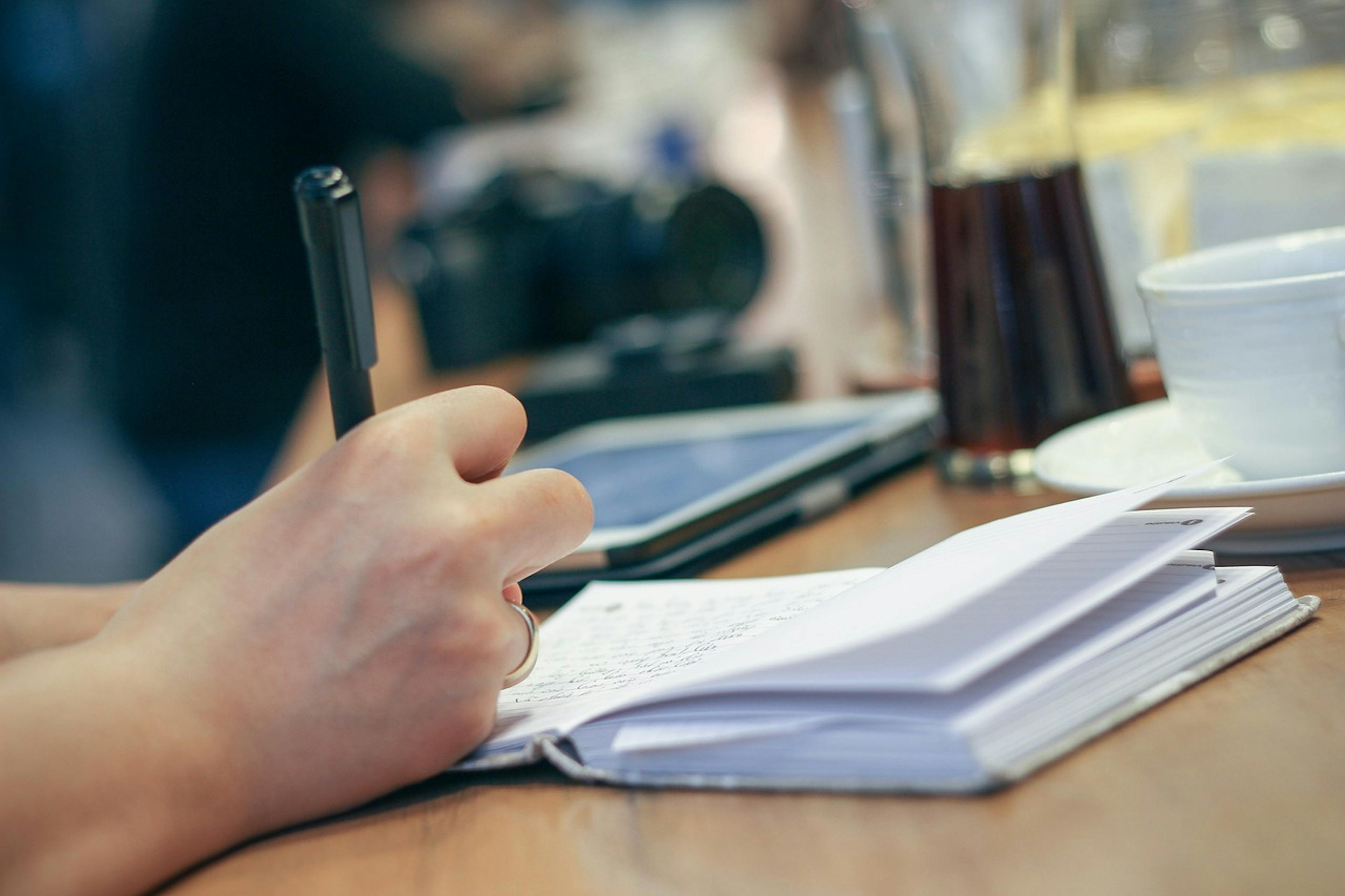 An image of someone writing in a notebook with a phone next to them - it is the main image for the article "Business Planner: 5 Proven Apps Used By Entrepreneurs"