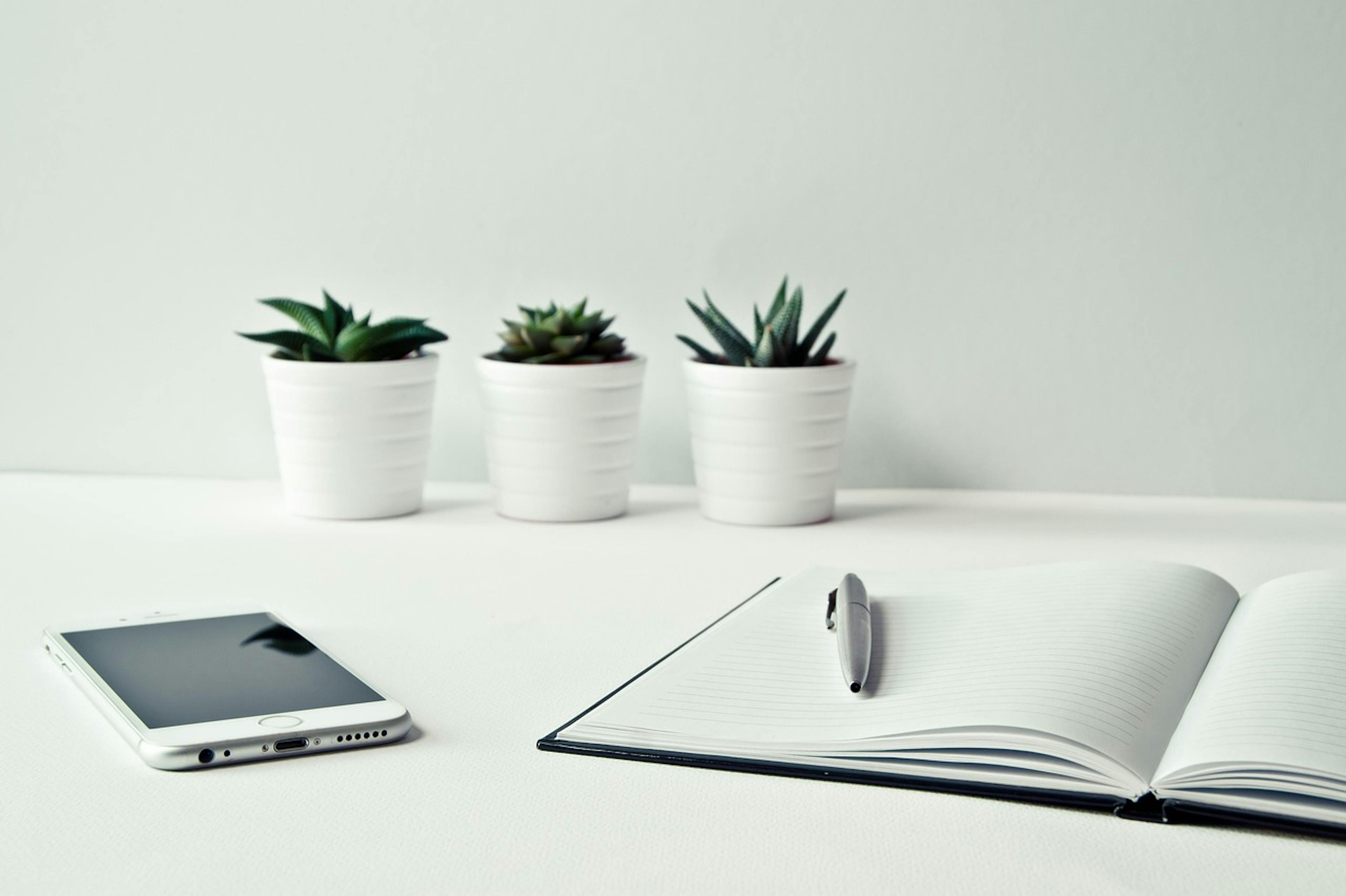 An image of a notebook and a phone on a table - it is the main image for the article "Self Care Journal: 6 Best Apps for Self-Care and Wellbeing"