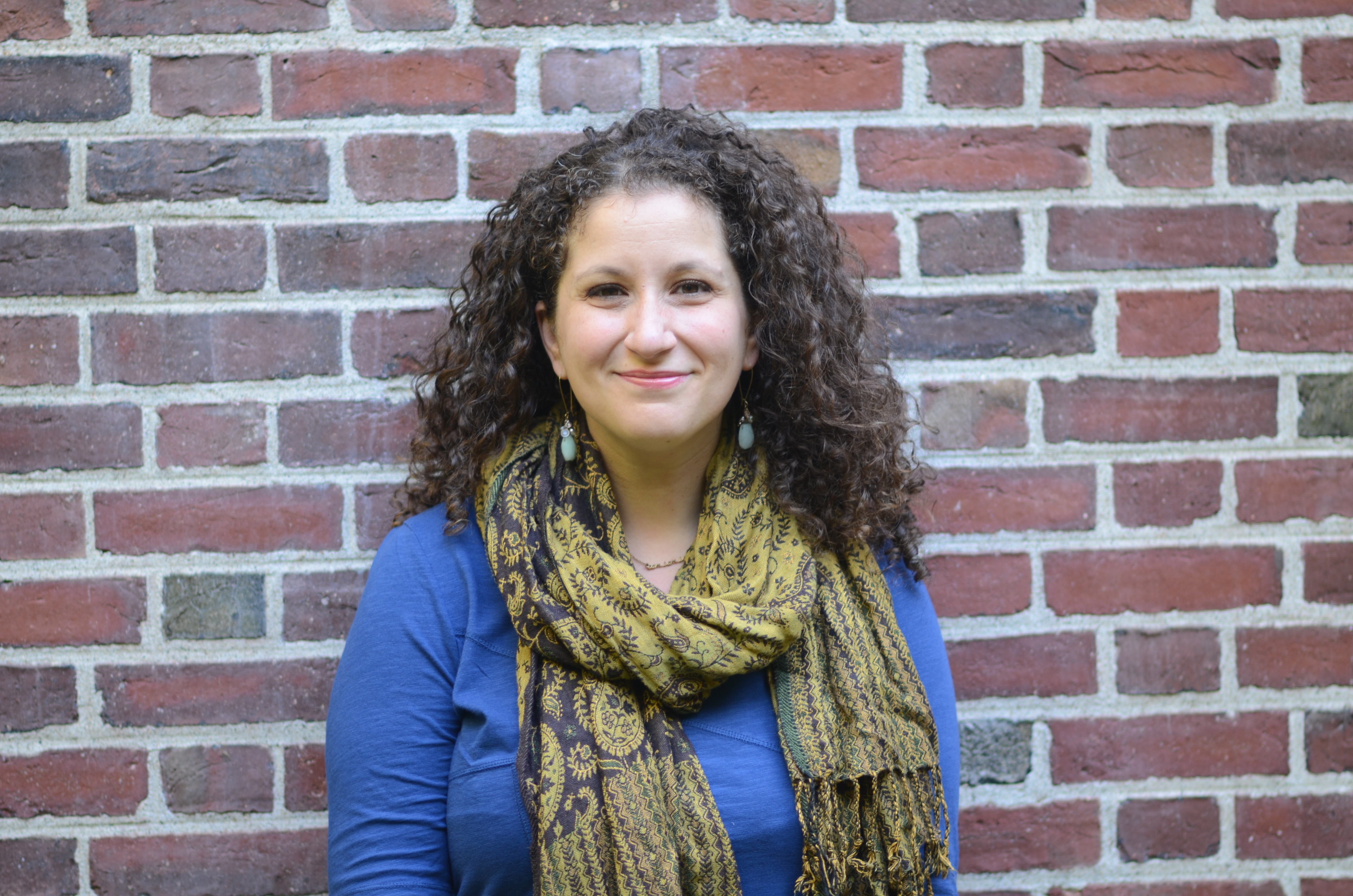 Vanessa Zoltan stands against a brick wall in a blue shirt and yellow scarf