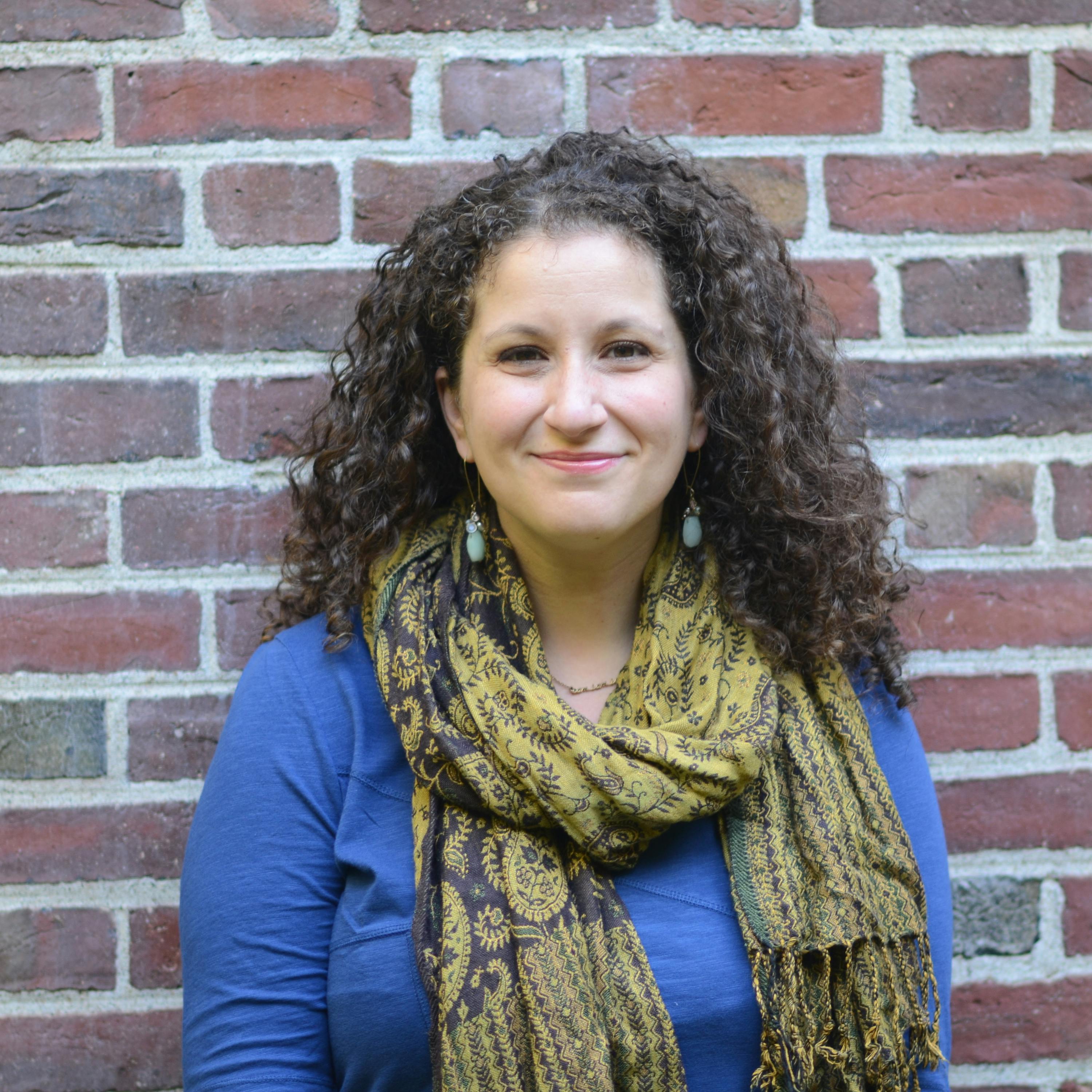 Vanessa Zoltan stands against a brick wall in a blue shirt and yellow scarf