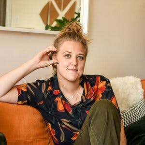 Profile photo of Hannah Rehak, sitting on an orange couch and wearing a shirt with red-orange flowers