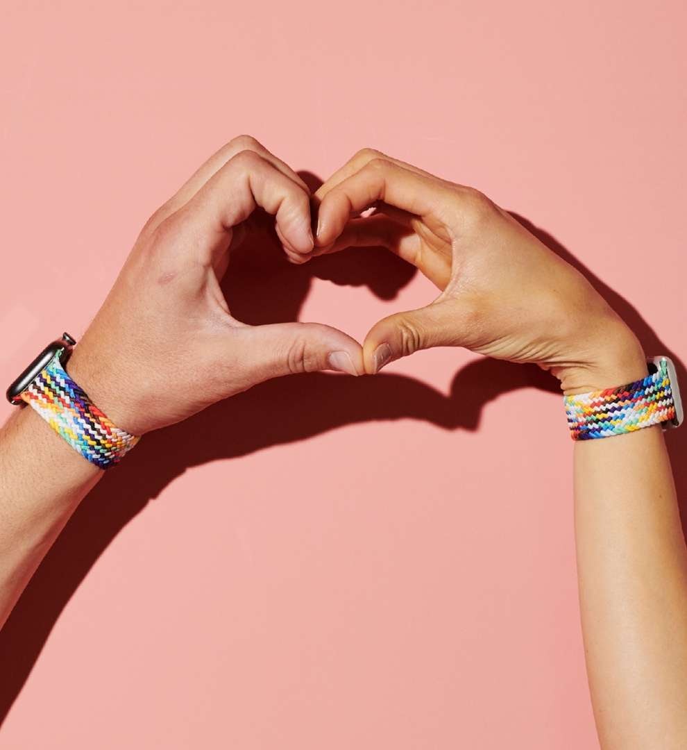 Image of two hands being held together to create a heart shape, both wearing Apple Watches© with multi-coloured straps.