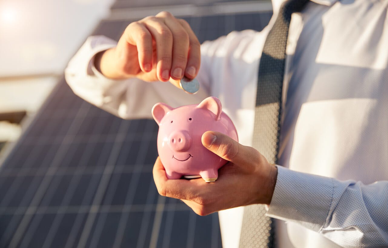 Homem perto de painel solar,  colocando dinheiro em cofrinho simbolizando economia