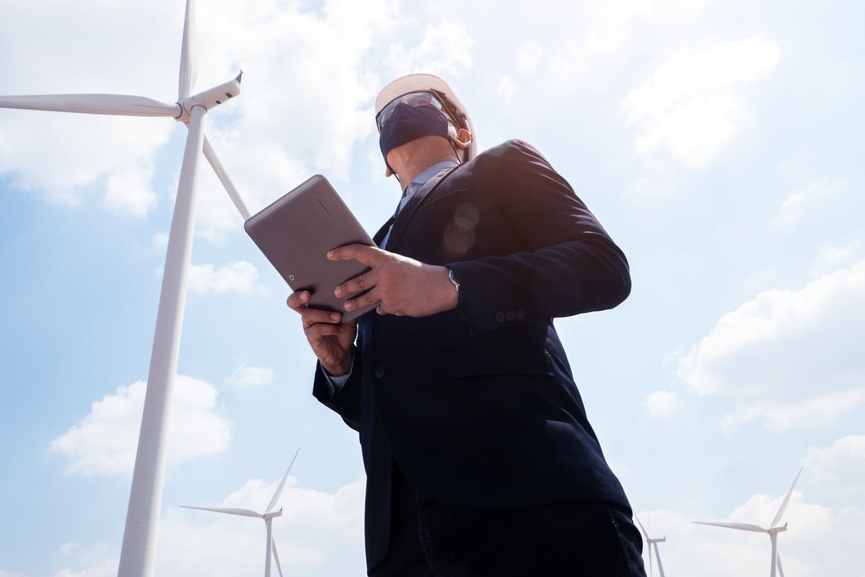Empresário de sucesso em moinhos de vento usando máscara facial e usando tablet com turbina eólica em segundo plano