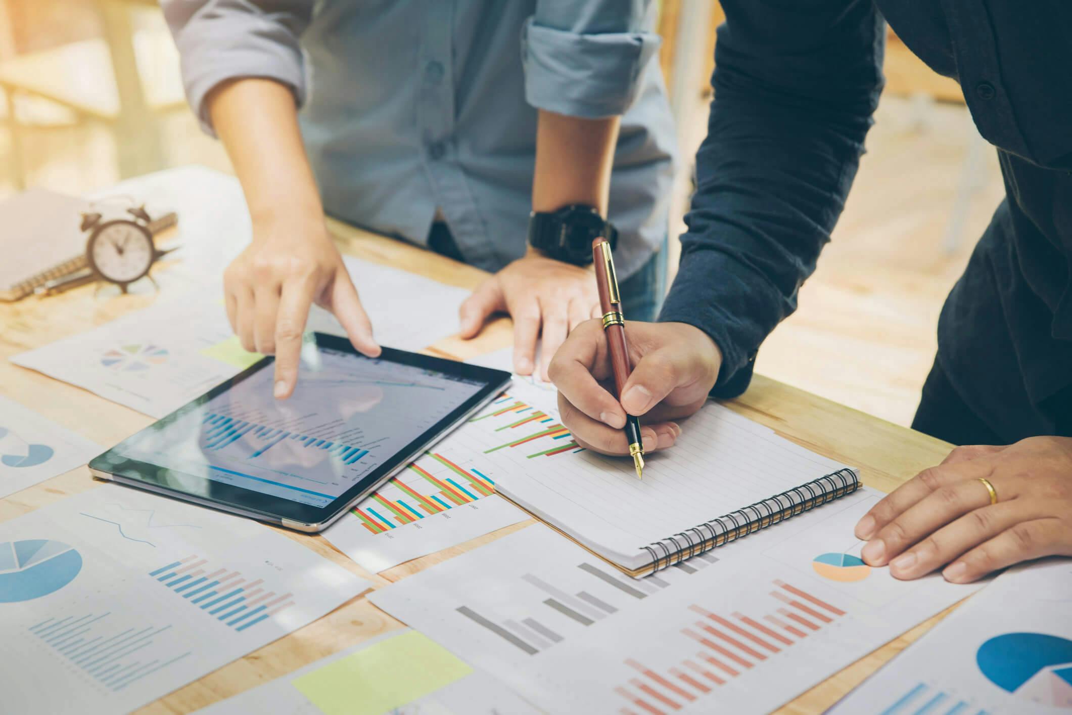 Dois homens analisando relatórios de dados e gráficos em cima de uma mesa de trabalho