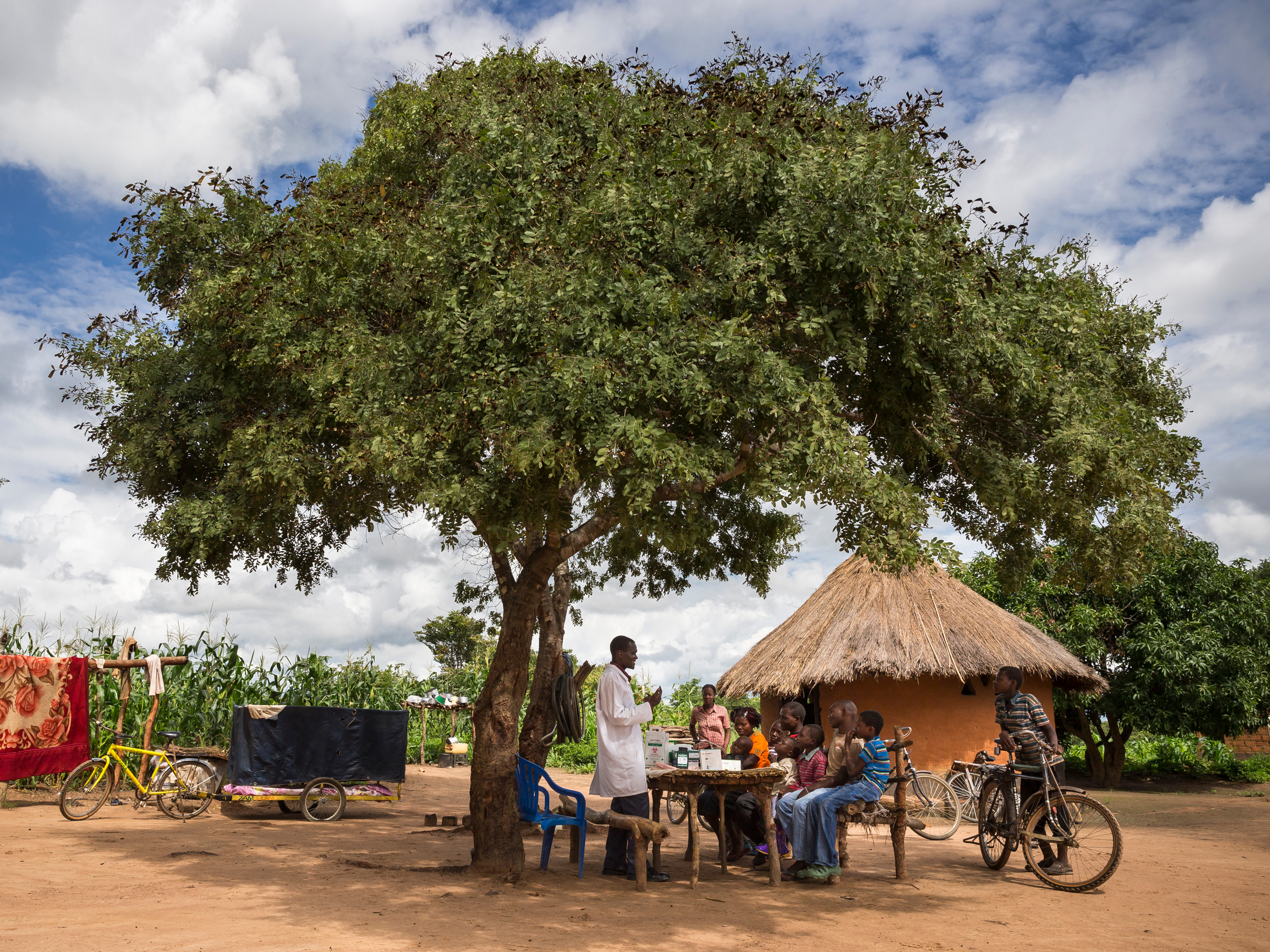 Ein Gesundheitshelfer mit einer Gruppe von Menschen, die unter einem großen Baum in ländlicher Umgebung um einen Tisch sitzen, mit einer strohgedeckten Hütte und Fahrrädern in der Nähe.