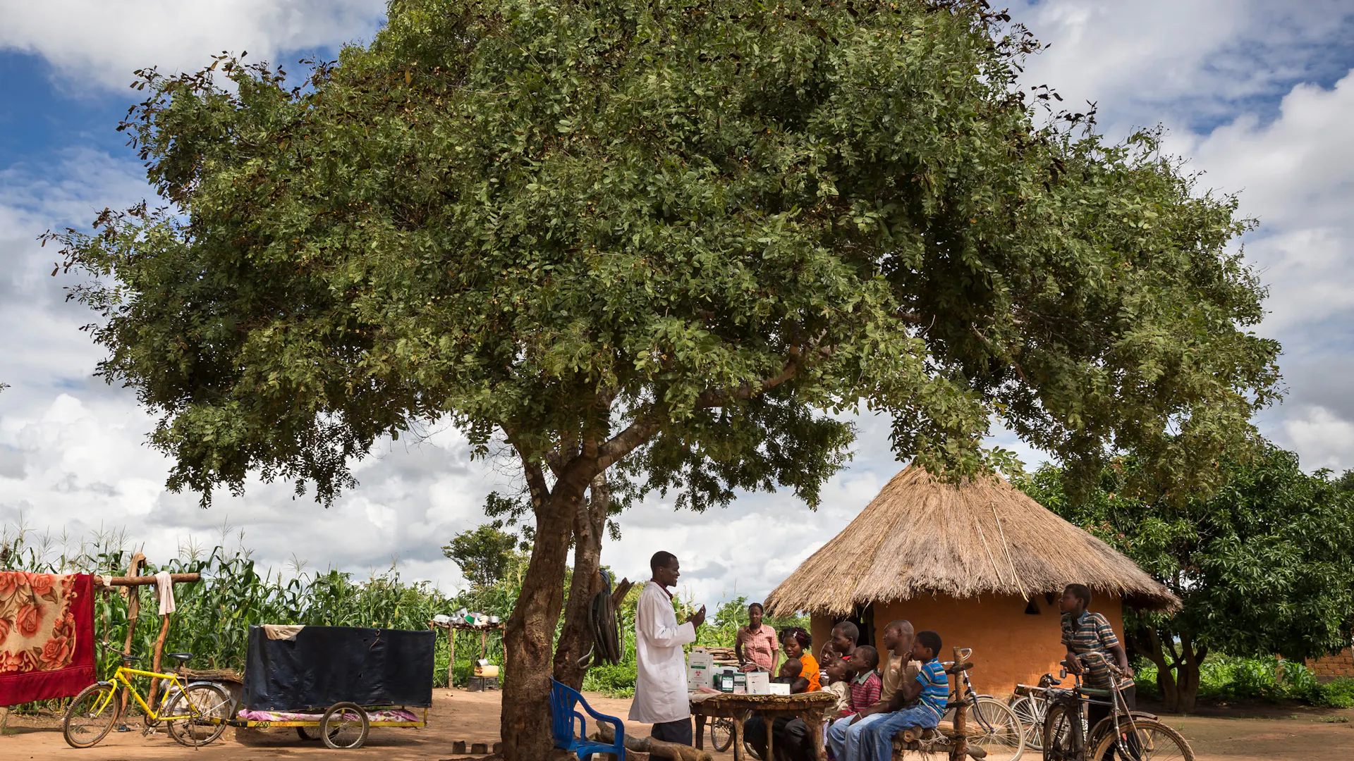 Ein Gesundheitshelfer mit einer Gruppe von Menschen, die unter einem großen Baum in ländlicher Umgebung um einen Tisch sitzen, mit einer strohgedeckten Hütte und Fahrrädern in der Nähe.