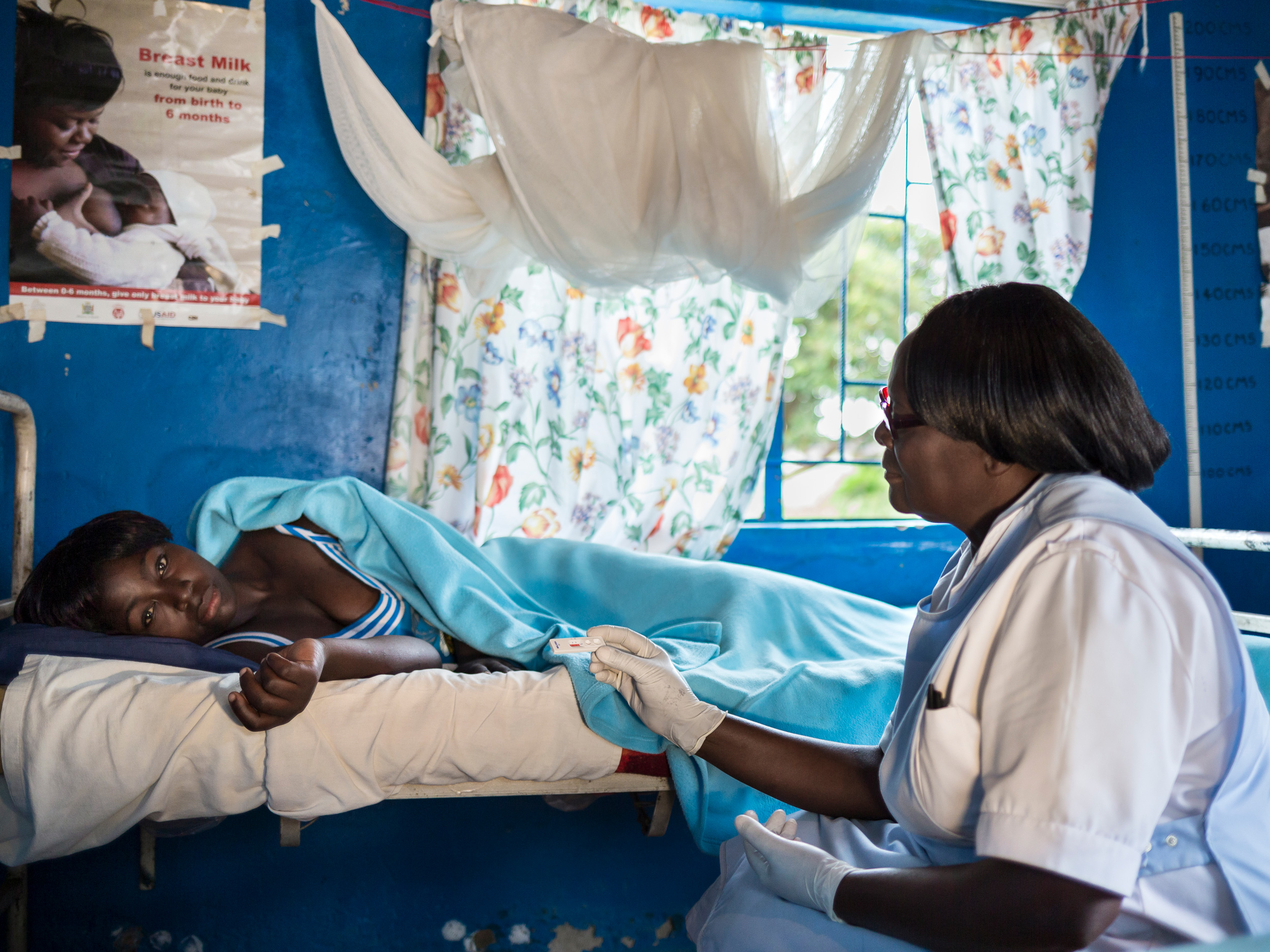 Eine medizinische Fachkraft in einem weißen Kittel sitzt neben einem Patienten, der auf einem Bett in einem Raum liegt, an dessen Wand ein Poster zum Thema Stillen hängt.