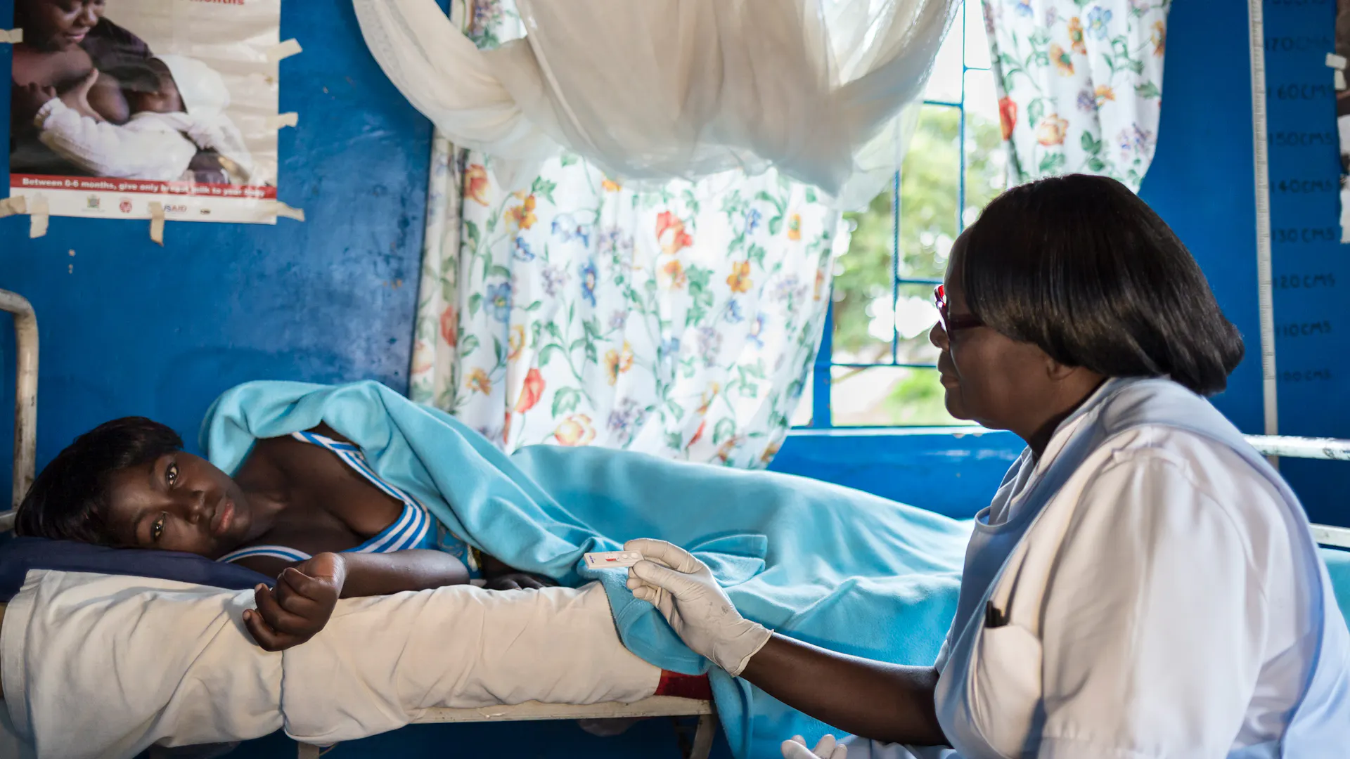 Eine medizinische Fachkraft in einem weißen Kittel sitzt neben einem Patienten, der auf einem Bett in einem Raum liegt, an dessen Wand ein Poster zum Thema Stillen hängt.